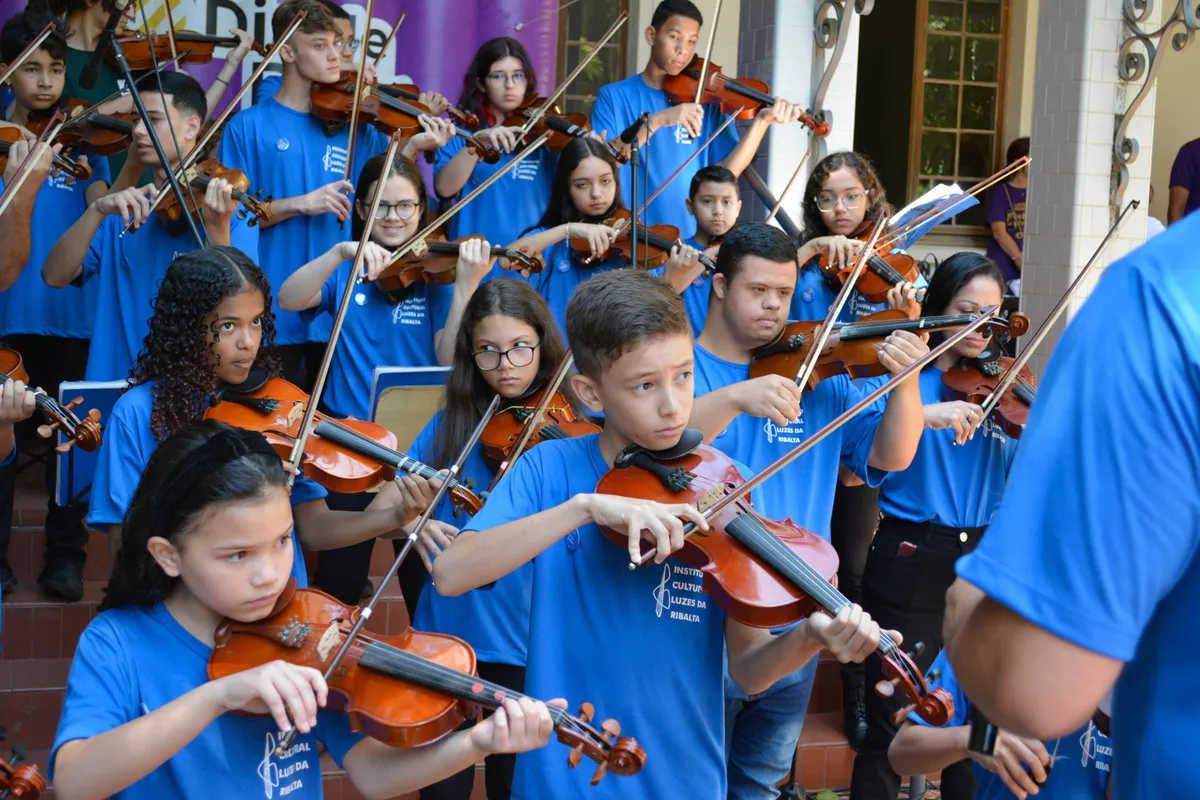 Alunos de camiseta azul do Instituto tocando violino ao ar livre, nos degraus de uma escadaria