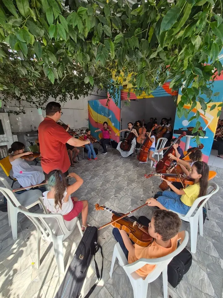 Professor orientando alunos de violino sentados em roda sob a parreira do pátio