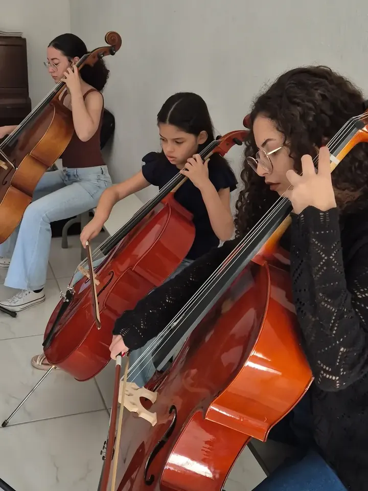 Três alunas tocando violoncelo lado a lado durante a aula