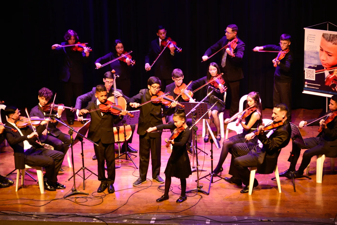 Orquestra Jovem em palco de teatro, com violinos, viola e violoncelo sob luzes coloridas